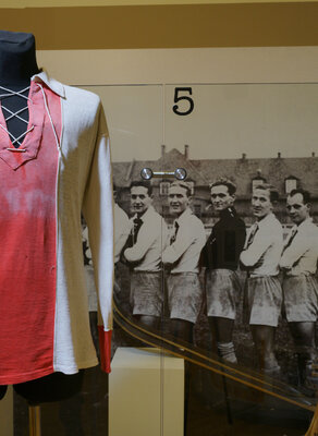 Zdjęcie przedstawia koszulkę piłkarską KS Gedania z długimi rękawami. Środek jest czerwony, boki i rękawy są białe. W tle czarno-białe zdjęcie przedstawiające sportowców z dwudziestolecia międzywojennego ustawionych w szeregu spoglądających na widza.   