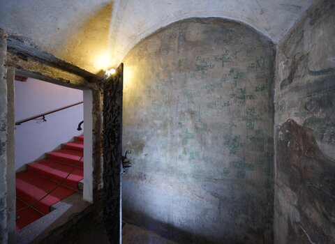 Zdjęcie przedstawia wnętrze pomieszczenia Mały Krzysztof. Po prawej otwarte drzwi i schody. Na ścianach wyblakłe malowidła z okresu średniowiecza przedstawiające mozaikę zielonych krzyży.  