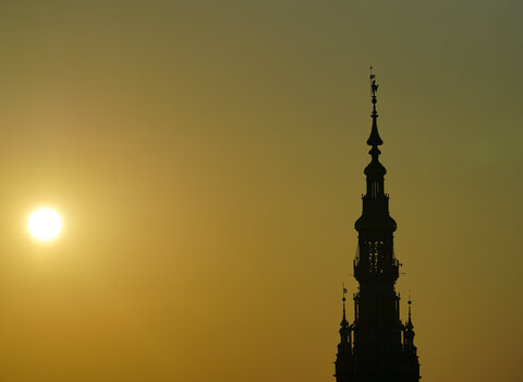 Zdjęcie przedstawia czarny szczyt spiczastej wieży Ratusza Głównego Miasta na tle złotego nieba. Po lewej zachodzące słońce. 