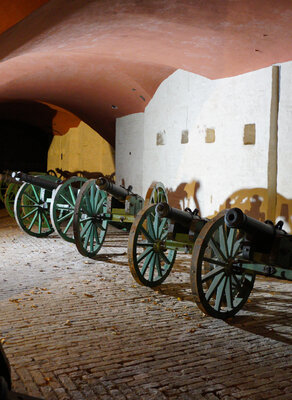 Zdjęcie przedstawia wnętrze bastionu Futra Wodna w Twierdzy Wisłoujście. Po prawej widać repliki czterech armat ustawionych pod ceglaną ścianą pomalowaną na biało. Sklepienie, podparte na filarach, jest w kolorze czerwonym.