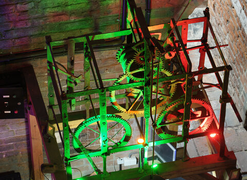 Zdjęcie przedstawia widok z górny na oświetlony światłami LED mechanizm starego zegara wieżowego. Konstrukcja szkieletowa, w środku koła zębate i przekładki. 