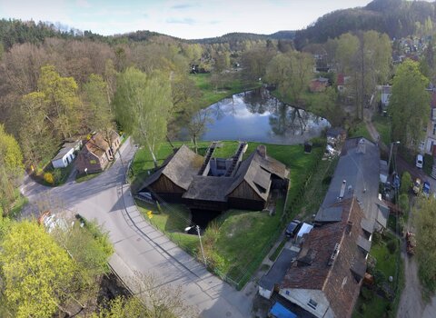 Zdjęcie wykonane z drona przedstawia budynek Kuźni Wodnej wraz z otoczeniem. W centralnym miejscu jest budynek kuźni, po lewej i prawej sąsiadujące budynki,  w tle zbiornik wodny na Potoku Oliwskim oraz Trójmiejski Park Krajobrazowy. 