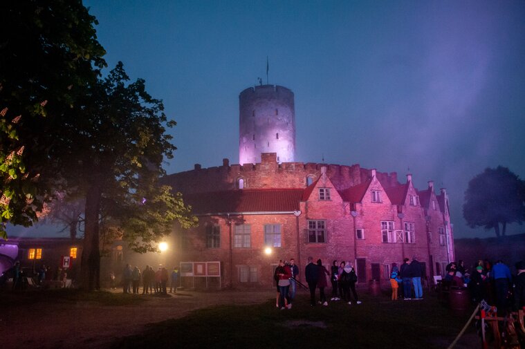 Zdjęcie przedstawia kamieniczki Wieńca Twierdzy Wisłoujście i dominującą nad nimi wieżę na tle wieczornego nieba. Kamieniczki są iluminowane światłem. Po lewej kasztan. Z prawej ludzie przy ognisku. 