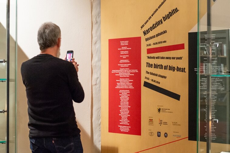 Zdjęcie przedstawia mężczyznę fotografującego planszę informacyjną o wystawie. Mężczyzna fotografujący wystawę telefonem znajduje się w lewej części zdjęcia (uchwycony od pasa w górę). Plansza wystawowa z dużym napisem "Nikt nam nie weźmie młodości. Narodziny bigbitu. Odsłona gdańska" i mniejszymi, nieczytelnymi.