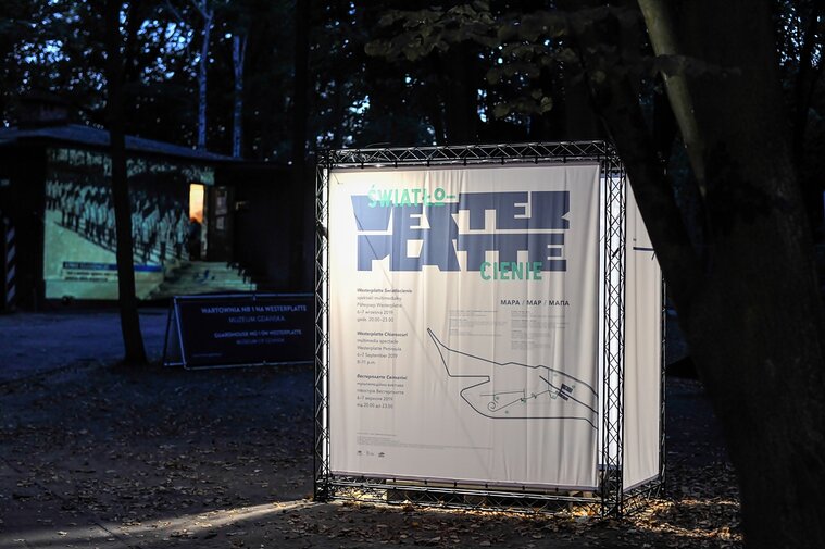 Zdjęcie przedstawia kubik informacyjny festiwalu "Westerplatte. Światłocienie". Bryła stoi na tle lasu. W tle prostopadłościenna bryła Wartowni nr 1 na Westerplatte. Na jej ścianie frontowej wyświetlany jest film.  