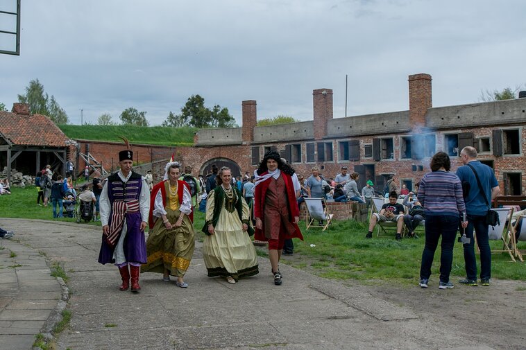 Majówka w Twierdzy Wisłoujście. Parada maszerujących przez plac rekonstruktorów i rekonstruktorek. Po prawej rząd turystów. W tle zabudowania koszar i wały. 