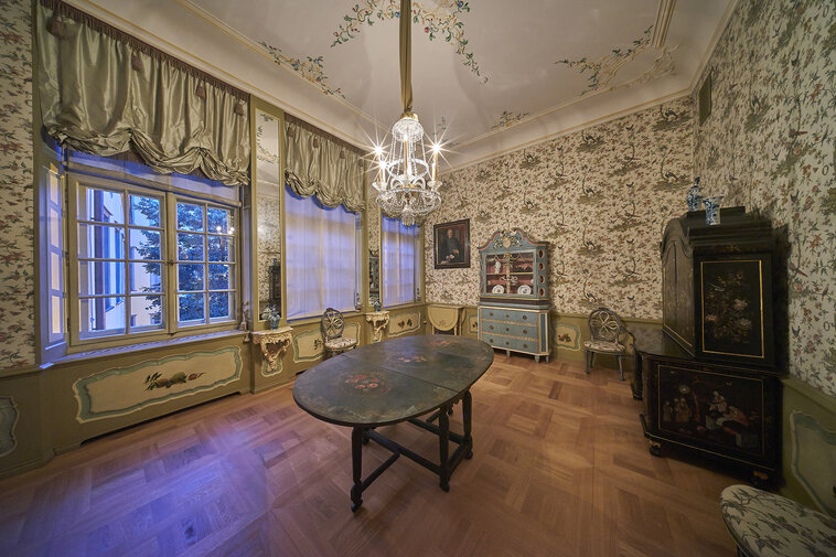 Zdjęcie przedstawia małą jadalnię. Na drewnianej podłodze stoi zdobiony owalny, składany stół, za nim kredens i meble, a po lewej stronie okno. Na ścianach znajdują się kremowe tapety zdobione motywami. Z białego sufitu zwisa lampa. 
