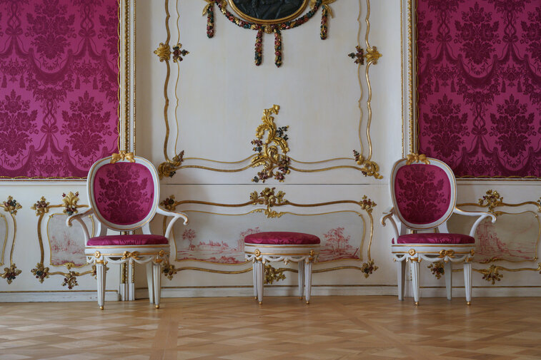 Zdjęcie przedstawia wnętrza Domu Uphagena. W centrum biała ściana, na prawo i lewo od niej szkarłatne, zdobione w formie smukłych ośmiokątów tapety. Na drewnianej podłodze stoją dwa białe krzesła obite szkarłatną tkaniną w kolorze tapet. Pomiędzy nimi niskie siedzisko obite tkaniną w tym samym kolorze.   