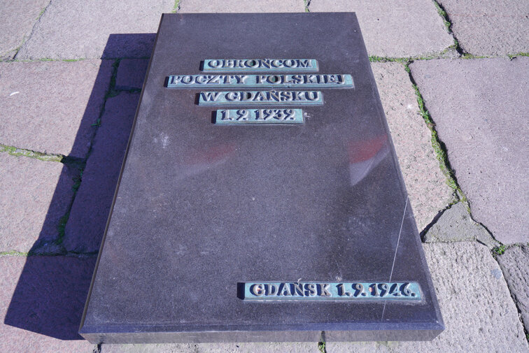 Zdjęcie przedstawia tablicę wmurowaną w posadzkę placu Obrońców Poczty Polskiej. Na kamieniu napis Obrońcom Poczty Polskiej w Gdańsku 1.9.1939 oraz Gdańsk 1.9.46