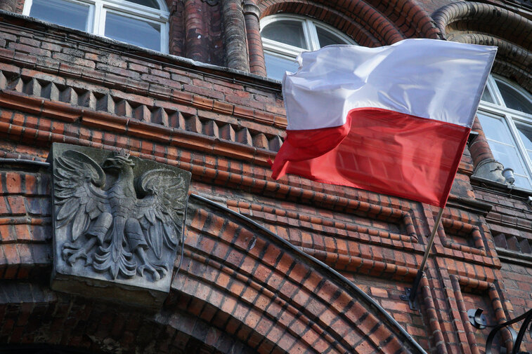 Zdjęcie przedstawia fragment fasady gmachu Poczty Polskiej. Nad drzwiami godło RP. Po prawej łopocze flaga Polski. 