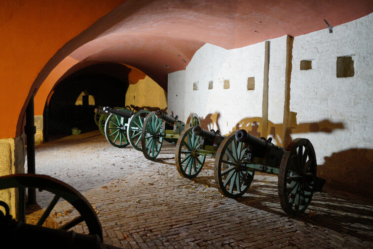 Zdjęcie przedstawia wnętrze bastionu Futra Wodna w Twierdzy Wisłoujście. Po prawej widać repliki czterech armat ustawionych pod ceglaną ścianą pomalowaną na biało. Sklepienie, podparte na filarach, jest w kolorze czerwonym.