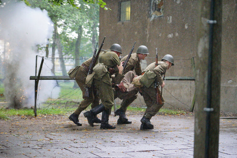 Zdjęcie przedstawia rekonstrukcję walk w obronie Westerplatte 1-7 września 1939 r. Widoczne sylwetki czterech żołnierzy niosących rannego kolegę do budynku wartowni. 