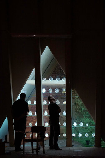 Zdjęcie przedstawia dwóch mężczyzn zwiedzających wystawę, patrzących na kolekcję zegarków stołowych ustawionych na oświetlonych półkach w kształcie piramidy. 