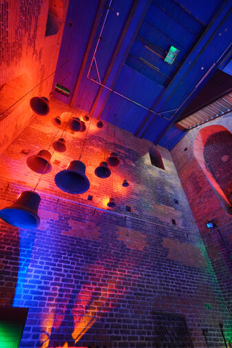 Zdjęcie przedstawia fragment sali wystawowej. Przy ceglanej ścianie zwisa z sufitu kilkanaście starych dzwonów oświetlonych czerwono-niebieskim światłem. 