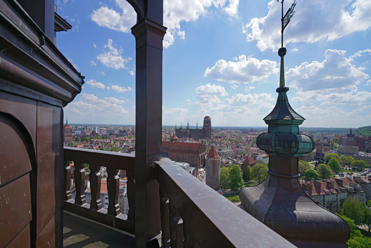 Zdjęcie panoramy Gdańska widocznej z wieży widokowej kościoła św. Katarzyny. Na pierwszym planie balustrada otaczająca punkt widokowy, w tle zabudowania Głównego Miasta z sylwetką Bazyliki Mariackiej. 