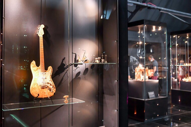 Zdjęcie. Fragment wystawy. Po lewej wisi gitara elektryczna wykonana z bursztynu bałtyckiego. Po prawej gabloty z bursztynowymi przedmiotami w tle.