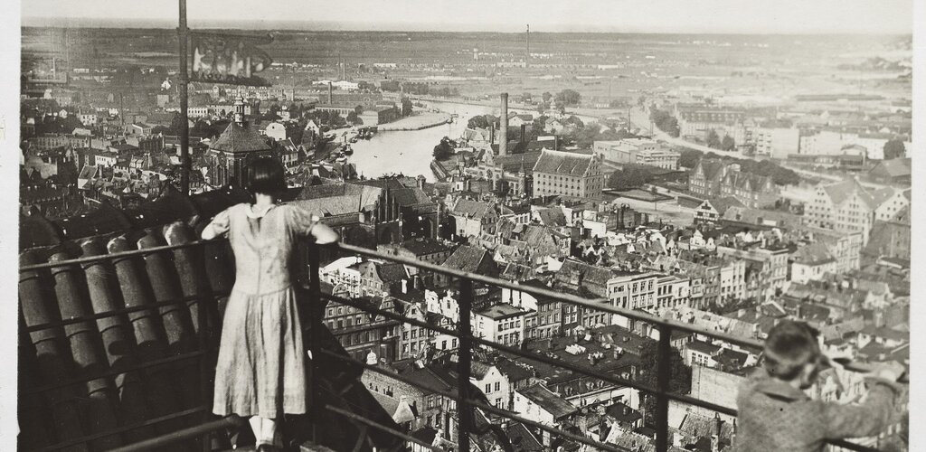 Zdjęcie przedstawia dziewczynkę na szycie wieży kościoła pod wezwaniem Najświętszej Maryi Panny. Postać uchwycona od tyłu spogląda na panoramę w kierunku Ołowiankę. W tle zabudowania, Motława, a dalej w głębi łąki.  