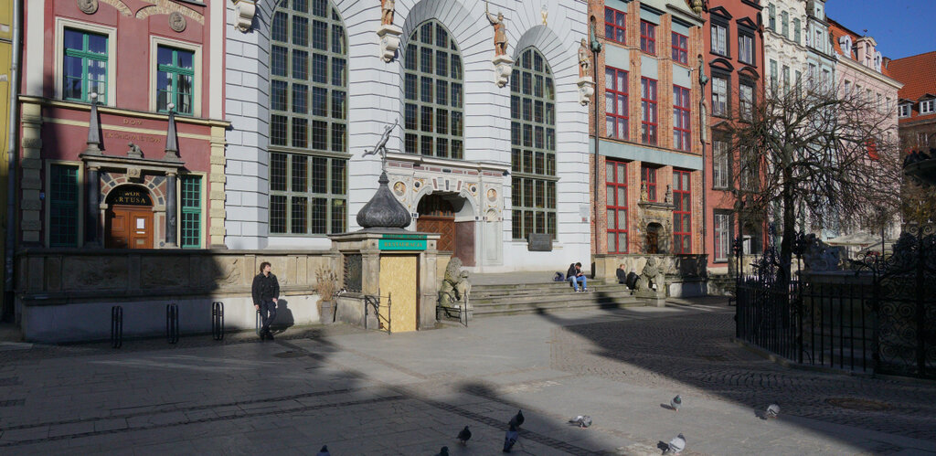 Fotografia ukazuje widok na Długi Targ w Gdański i białą fasadę Dworu Artusa z trzema wielkimi oknami oraz długim płaskim szczytem z balkonem. W tle sąsiadujące kamienice. W dolnej części zdjecia fragment ułożonego z płyt chodnika Długiego Targu.  