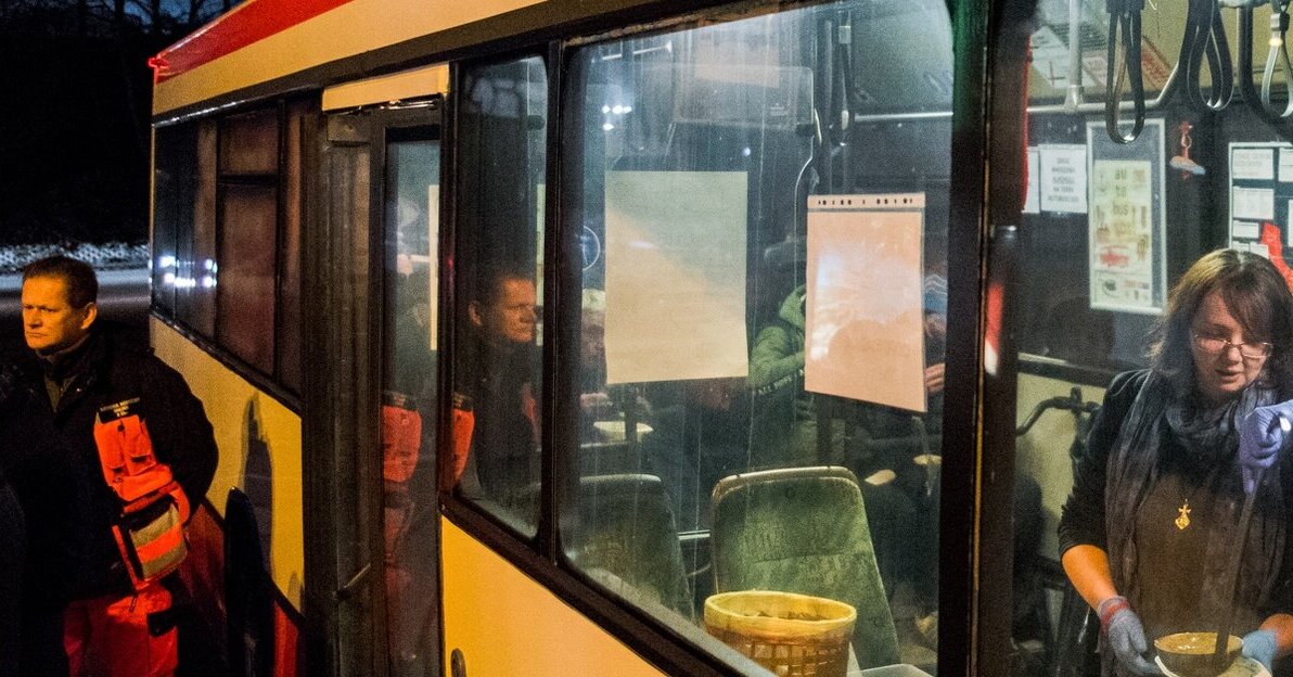 Na zdjęciu widoczny jest autobus, w środku znajduje się kobieta nalewająca zupę, przed wejściem do pojazdu stoi ratownik medyczny.