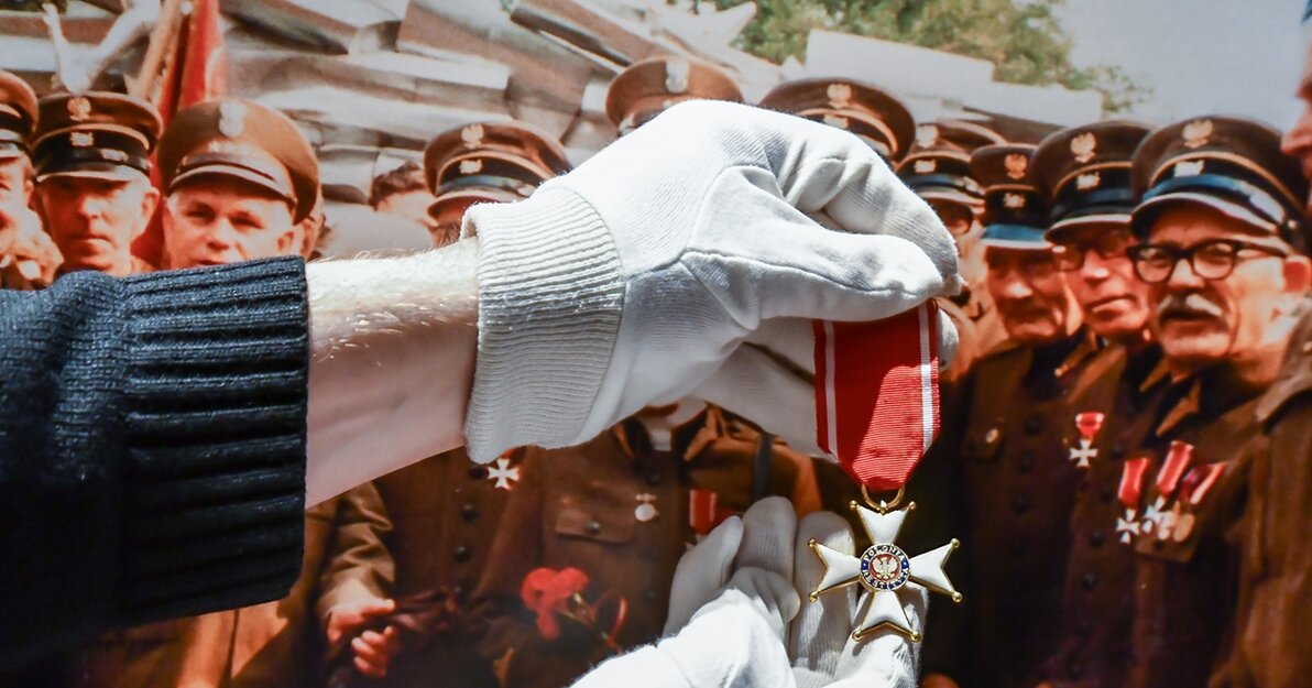 Zdjęcie przedstawia dłonie w rękawiczkach trzymające Order Polonia Restituta. W tle zdjęcie z uroczystości. Widnieją na nim starsi mężczyźni ubrani w mundury. Na piersiach mają odznaczenia. Order trzymany w dłoniach jest podobny do noszonego na mundurze jednego z członków uroczystości, Alfonsa Pillatha (po prawej od dłoni).   