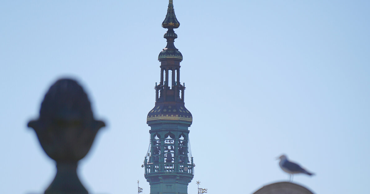 Gdańsk. Wieża Ratusza Głównego Miasta. Poniżej niej fragmenty dwóch szczytów gdańskich kamienic. Na prawej siedzi ptak.   