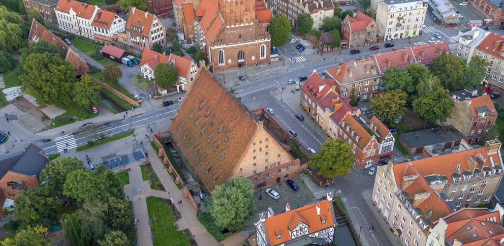 Zdjęcie przedstawia widok na ulice Starego Przedmieścia z lotu ptaka. W centrum znajduje się bryła Wielkiego Młyna ze spadzistym dachem i opływający go Kanał Raduni. Nieco wyżej ulica Wielkie Młyny oraz kościół św. Katarzyny wraz z wieżą kościoła. Poniżej zielone drzewa i Skwer Heweliusza.  