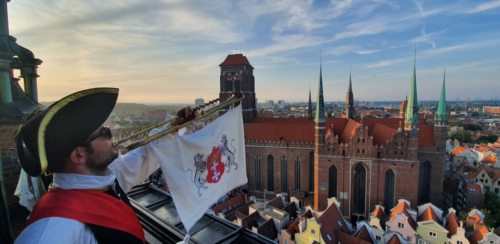 Zdjęcie przedstawia trębacza ubranego w historyczny strój. Do trąbki przymocowany chwytak, na nim flaga a na niej z lwy trzymające herb Gdańska. W tle zabudowania oraz Bazylika Mariacka.  