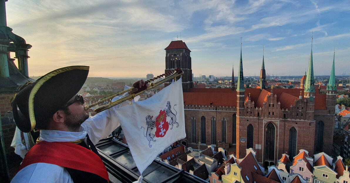 Zdjęcie przedstawia trębacza ubranego w historyczny strój. Do trąbki przymocowany chwytak, na nim flaga a na niej z lwy trzymające herb Gdańska. W tle zabudowania oraz Bazylika Mariacka.  