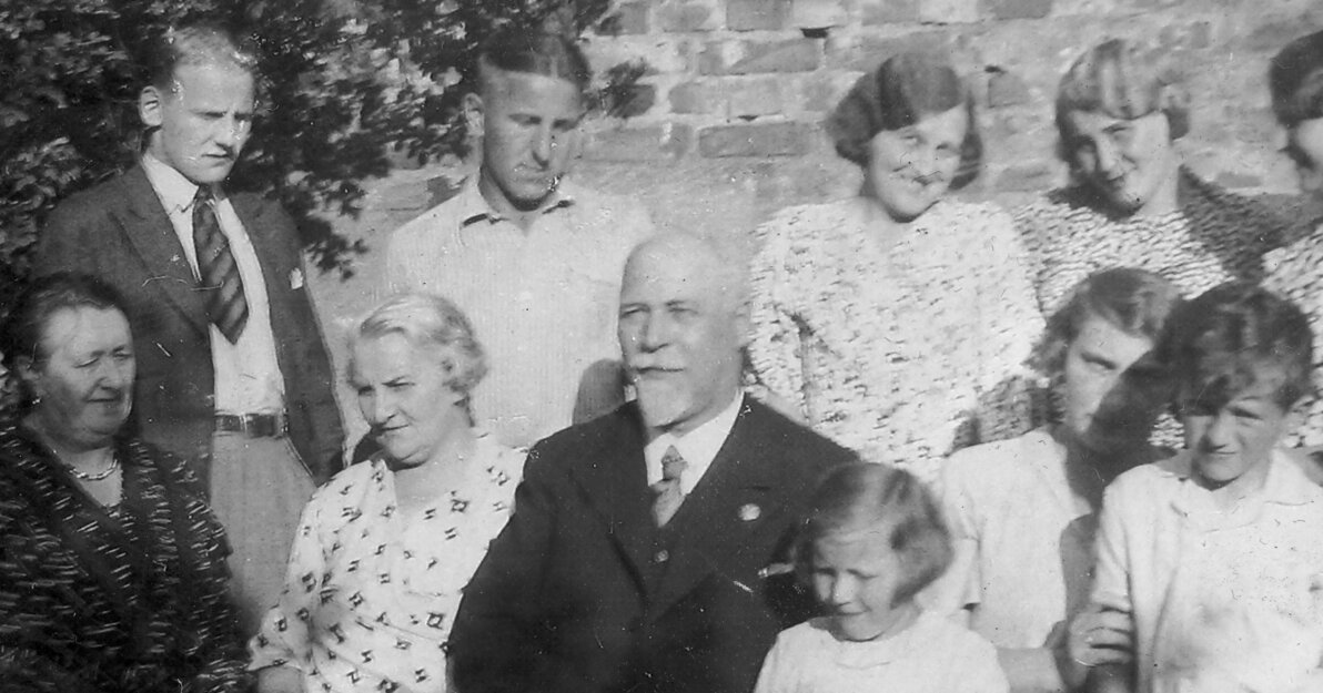 Zdjęcie przedstawia rodzinę Bellwonów - 11 osób. W centrum starszy mężczyzna i kobieta otoczone przez synów, córki i dwoje wnucząt. Za nimi mur. Po lewej stronie fragment drzewa. 