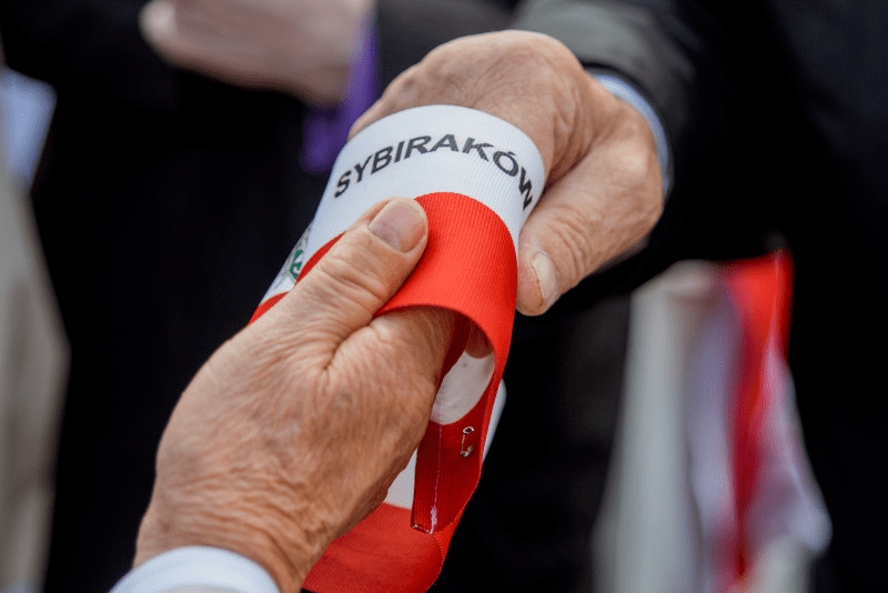 Na zdjęciu znajdują się dłonie starszych mężczyzn, które podają sobie biało-czerwoną opaskę naramienną z napisem Sybiracy. 