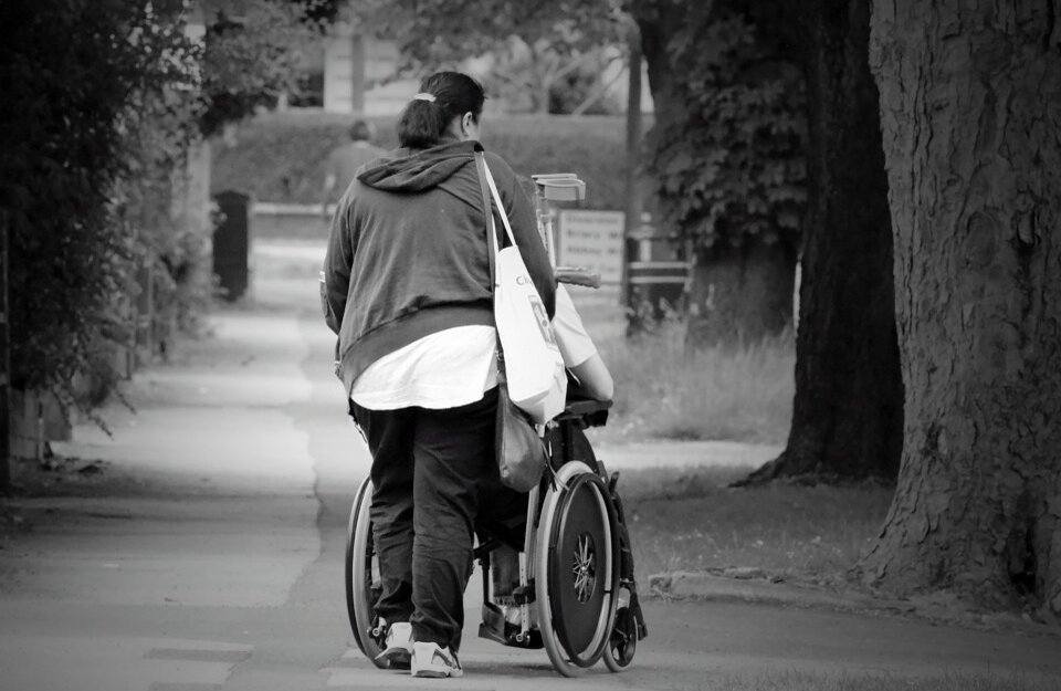 Na zdjęciu przedstawiono osobę poruszająca się na wózku i druga osobę, która ten wózek prowadzi. Osoby te znajdują się na alejce w parku. 