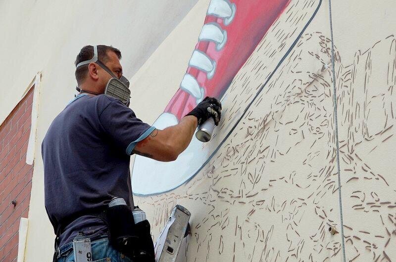 Zdjęcie przedstawia Looneya w trakcie tworzenie pierwszego muralu w ramach akcji Gdańsk bez plastiku. Koloruje on sprayem szkic buta znajdujący się na murze. Na twarzy ma maskę zabezpieczającą przed wdychaniem szkodliwych oparów sprayu. 