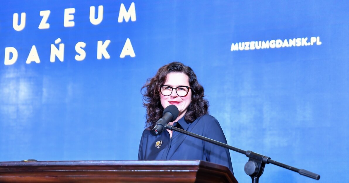 Zdjęcie przedstawia prezydent Gdańska, Aleksandrę Dulkiewicz przemawiającą z mównicy na tle ekrany z napisem 50 lat Muzeum Gdańska.  