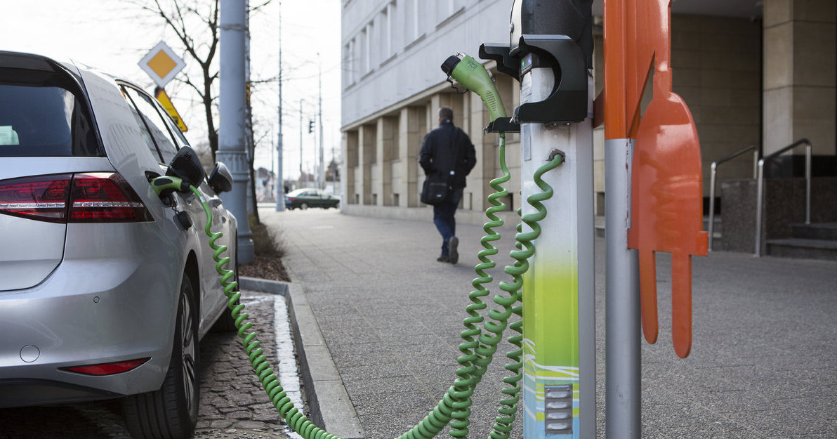 Na zdjęciu widoczna jest ładowarka do samochodu elektrycznego, z której ładowany jest pojazd. Ładowarka znajduje się na chodniku, w tle widać budynek.