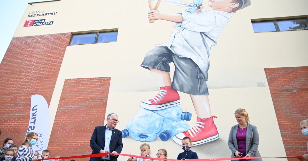 W tle zdjęcia znajduje się mural przedstawiający chłopca, który z procy wystrzela plastikową butelkę - pozbywa się plastiku. Na pierwszym planie prezydent Piotr Kowalczuk wraz z uczniami szkoły przecina czerwoną wstęgę z herbem Gdańska - symbolicznie odsłania mural. 