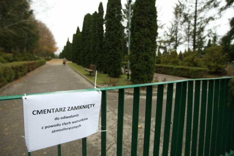 Na zdjęciu widoczne zielone ogrodzenie, w tle aleja cmentarza, z rzędem drzew iglastych po prawej stronie. Na ogrodzeniu biała tablica z informacją, że  z powodu złych warunków atmosferycznych cmentarz zamknięty do odwołania.