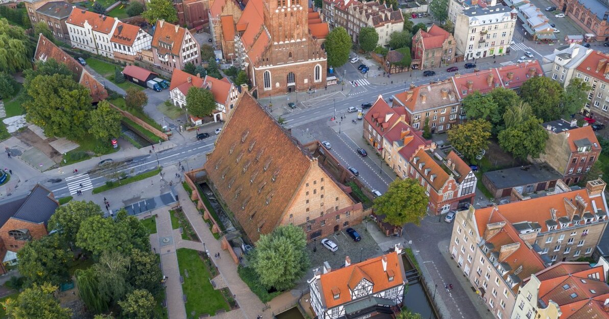 Zdjęcie przedstawia widok na ulice Starego Przedmieścia z lotu ptaka. W centrum znajduje się bryła Wielkiego Młyna ze spadzistym dachem i opływający go Kanał Raduni. Nieco wyżej ulica Wielkie Młyny oraz kościół św. Katarzyny wraz z wieżą kościoła. Poniżej zielone drzewa i Skwer Heweliusza.  