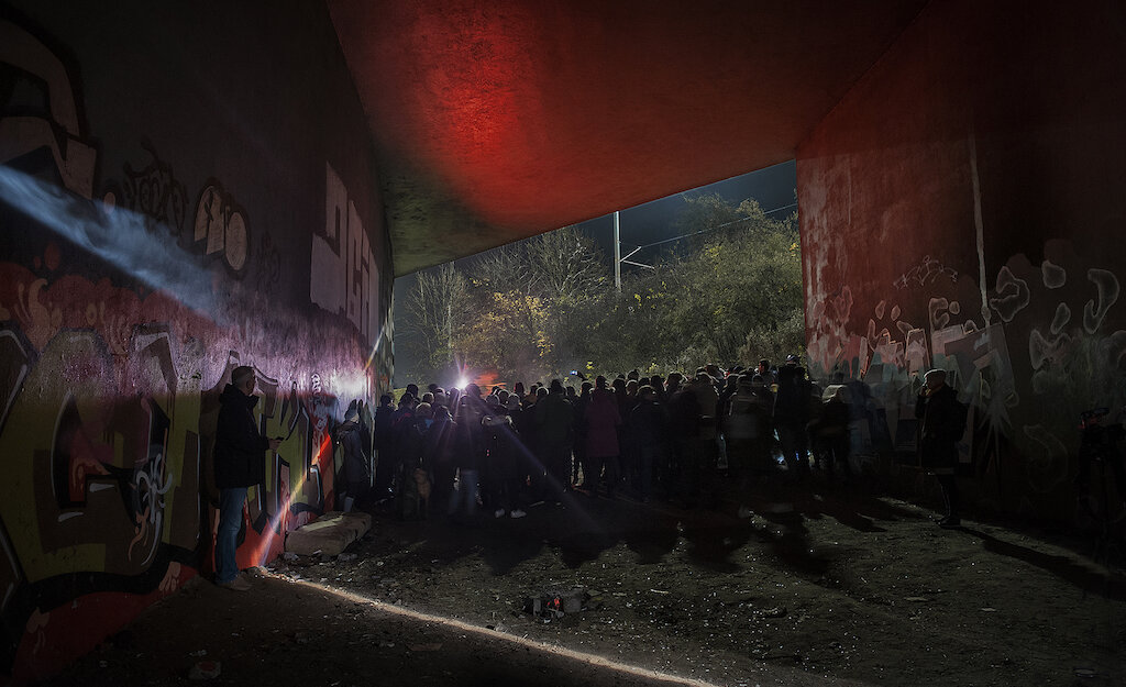 Zdjęcie zrobione nocą podczas 11 edycji Narracji. Tłum ludzi stoi pod wiaduktem, w tunelu, na murach jest graffiti Wszystko oświetlone jest na czerwono, prześwitują refleksy białego światła. 