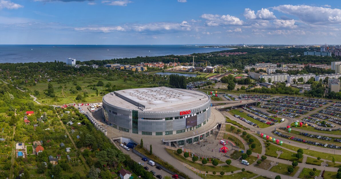 Panorama z widokiem na Ergo Arenę. Zdjęcie zrobione z dużej wysokości. W centralnej części zdjęcia okrągła, srebrna hala. W bezpośrednim sąsiedztwie hali parkingi oraz tereny zielone. Z prawej strony widać wysokie zabudowania Żabianki. W tle wody Zatoki Gdańskiej. 