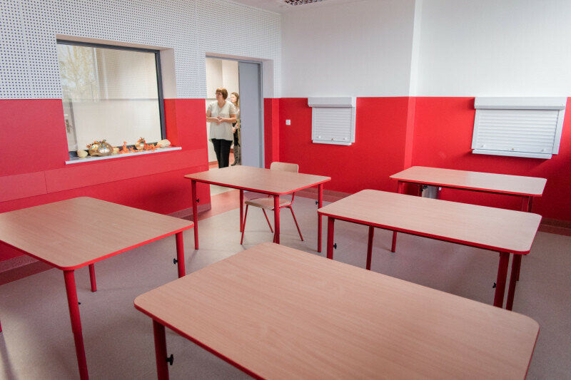 Na zdjęciu pusta klasa szkolna. W sali same ławki i krzesła. Otwarte drzwi do klasy, w których widać sylwetkę nauczyciela.