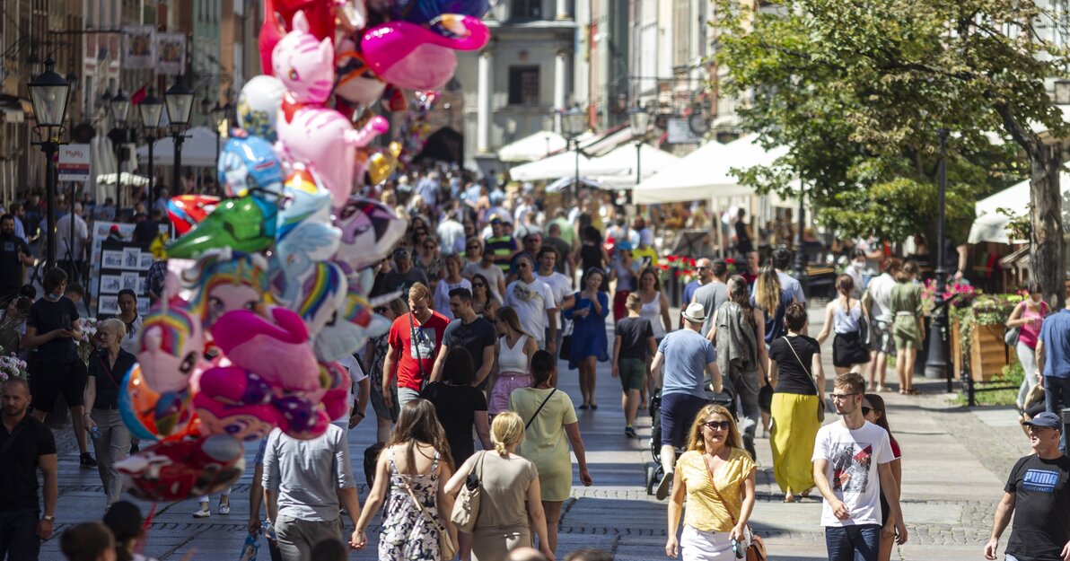 Wypełniona turystami ul. Długa w Gdańsku.  Z tłumu turystów wyróżnia się kobieta prowadząca psa na smyczy. Po lewej stronie zdjęcia, nieco nieostry pęk kolorowych balonów. W tle widać Złotą Bramę. 