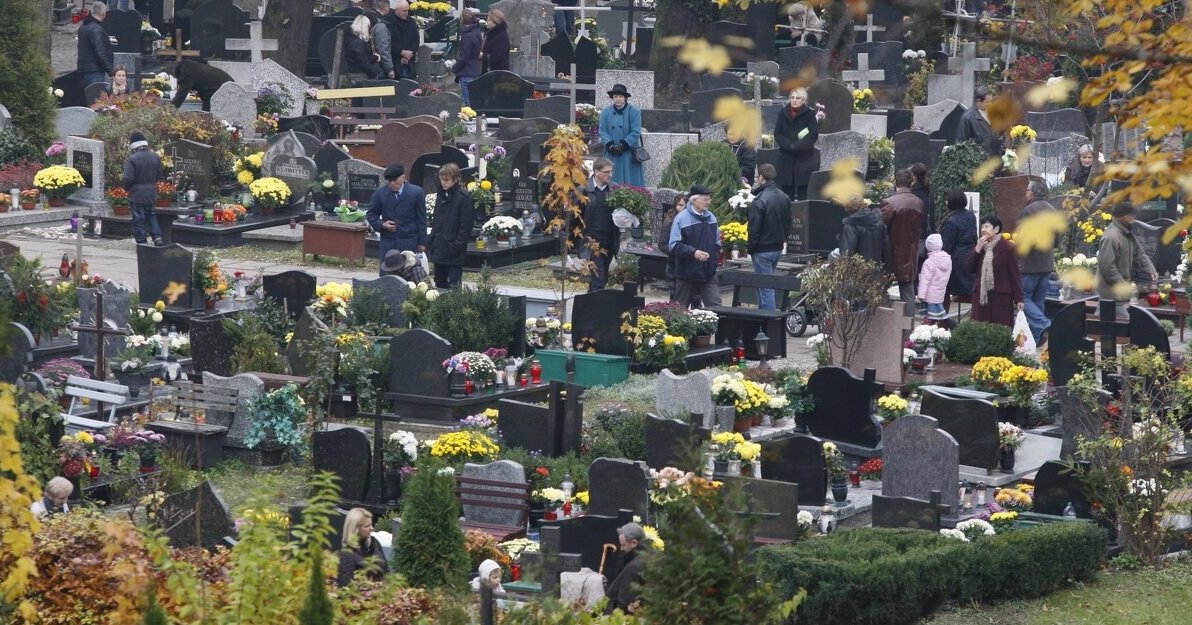 Zdjęcie przedstawia widok ze wzniesienia na Cmentarz Garnizonowy. Widoczne są nagrobki oraz kilka postaci stojących przy pomnikach. Pomiędzy grobami widoczne drzewka w kolorach jesieni zółtym, pomarańczowym, brązowym