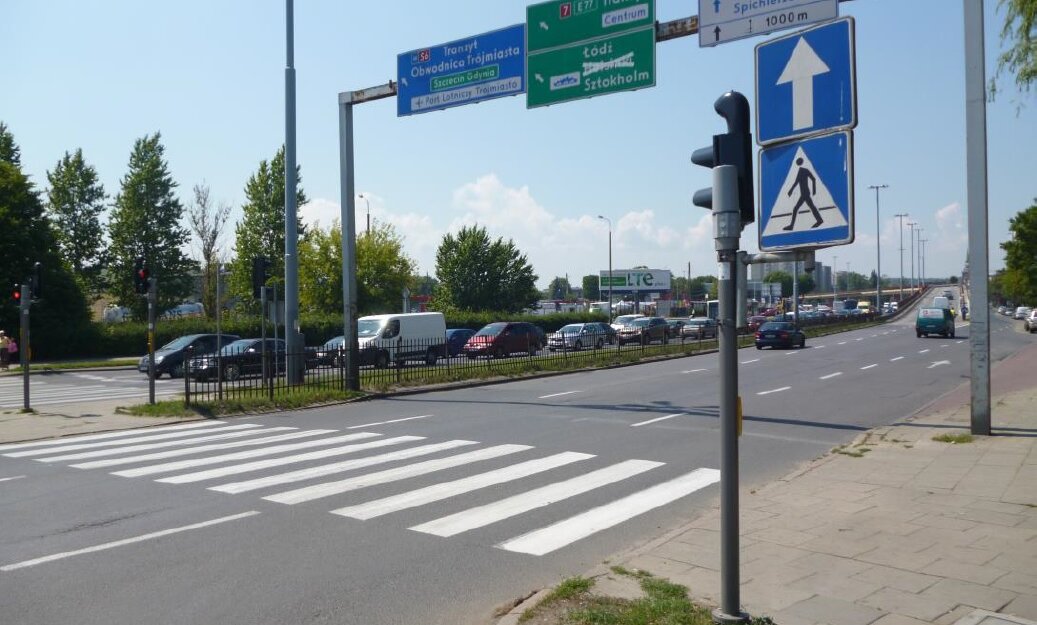 Na zdjęciu widoczna jest szara kilkupasmowa jezdnia, a nad nią stoi bramownica, czyli konstrukcja ponad jezdnią na której zawieszone są znaki drogowe. Widoczne są też znaki drogowe i samochody.