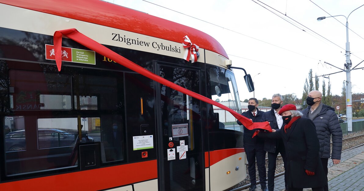 Czterech mężczyzn, odesłania umieszczony na burcie tramwaju napis - Zbigniew Cybulski. Panowie, którzy zaciągają czerwoną wstęgę są ubrani na ciemno i mają założone maseczki. Jeden z nich - radny Andrzej Stelmasiewicz ma czerwone buty, rękawiczki, szalik i beret. 