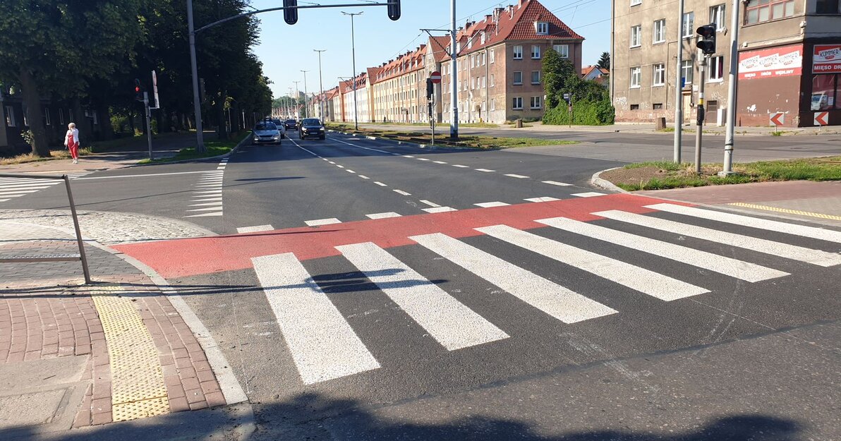 Zdjęcie przedstawia skrzyżowanie przy al. Hallera w Gdańsku. Na pierwszym planie białe pasy na przejściu dla pieszych oraz czerwona ścieżka rowerowa przecinająca ulicę. Al. Hallera ma po 3 pasy ruchu w obu kierunkach. Po lewej stronie, nieco w głębi kilka samochodów czeka na zmianę światła. Po prawej stronie widać czteropiętrowe kamienice ze spadzistymi dachami, po lewej szpaler wysokich drzew otaczających chodnik.   