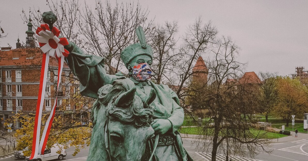 Pomnik Jana II Sobieskiego na koniu z kotylionem w ręku. Zdjęcie przedstawia pomnik polskiego króla, który siedzi na koniu. Jego jedna ręka jest wyciągnięta, przypięty do niej jest białoczerwony kotylion ze zwisającymi wstęgami. Zdjęcie zrobione jest z pewnej wysokości. W tle widać drzewa, ulice i zabudowania śródmieścia Gdańska. Monument ma kolor zielony. Król Sobieski ma założoną kolorową maseczkę.  