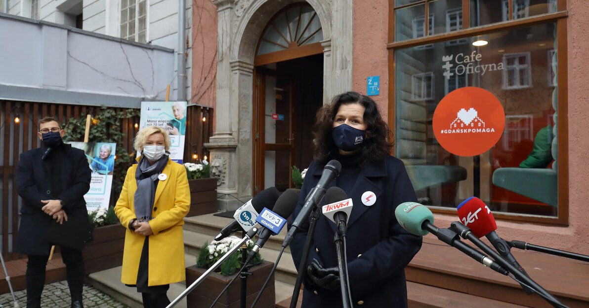 Zdjęcie zrobione podczas konferencji, Alekstandra Dulkiewicz, prezydent Gdańska mówi do mikrofonów, obok stoją Paweł Buczyński, Wiceprezes Fundacji Gdańskiej i Małgorzata Niemkiewicz, dyrektor Miejskiego Ośrodka Pomocy Rodzinie w Gdańsku. Stoją przed witryną Cafe Oficyna, na której znajduje się czerwona, okrągła naklejka Gdańsk pomaga. 