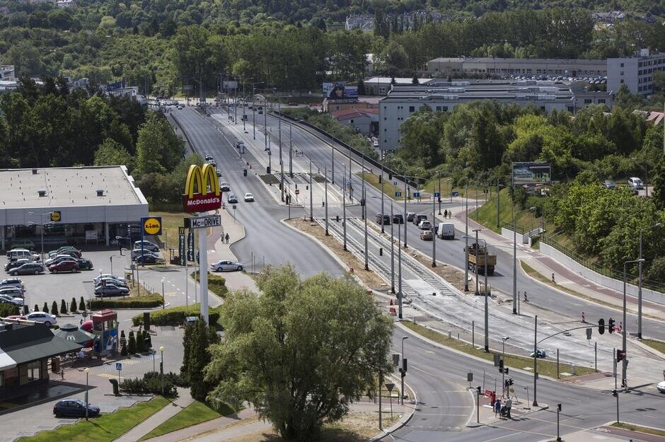 Zdjęcie przedstawia widok z góry na szeroką, dwujezdniową drogę. W każdym kierunku po 3 pasy ruchu. Między jezdniami tory tramwajowe i słupy trakcyjne. Po prawej stronie drzewa oraz zabudowania. W centralnej, górnej części zdjęcia oddalone drzewa. Po lewej stronie sklep popularnej, niemieckiej marki oraz restauracja z hamburgerami jeszcze popularniejszej, amerykańskiej marki.   