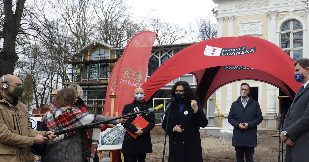 Na zdjęciu prezydent Dulkiewicz, zastępcy i radni gdańska, podczas konferencji prasowej. Za nimi widoczny namiot w barwach Gdańska, a w tle remontowany Dom Zdrojowy.