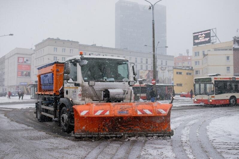 Pomarańczowy samochód typu pług śnieżny, jedzie zaśnieżoną ulicą. W tle po prawej stronie widać stojący autobus. Pług zgarnia śnieg przed sobą. Ale nie są to duże ilości śniegu. Fot. D. Paszliński